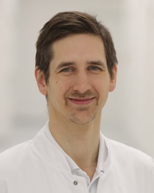 Ein Mann in einem weißen Kittel steht in einem hellen Flur. Er hat dunkles Haar und lächelt leicht. Der Hintergrund ist unscharf und zeigt eine moderne, klinische Umgebung.