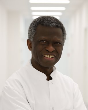 Ein lächelnder Arzt mit grauen Haaren trägt einen weißen Kittel und steht mit verschränkten Armen in einem hellen Krankenhausflur.