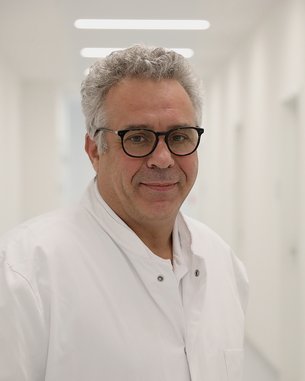 PD Dr. med. Matthias Goos Ein Arzt in einem weißen Kittel steht in einem hellen Flur. Er trägt eine Brille und lächelt leicht.