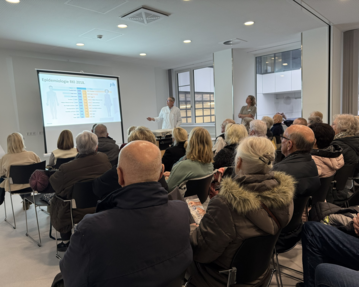 Ein Referent präsentiert Informationen zu Epidemiologie vor einem Publikum in einem modernen Raum. Die Zuhörer sind aufmerksam und sitzen in Reihen.