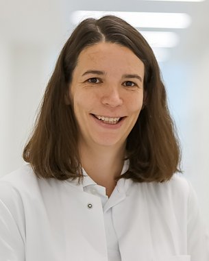 Eine Frau mit langen, braunen Haaren trägt einen weißen Kittel und lächelt freundlich. Der Hintergrund ist hell und unscharf, was den Fokus auf die Person lenkt.