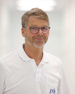 Ein Mann mit Brille und grauen Haaren trägt ein weißes Polohemd mit einer Brusttasche, auf der "srh" steht. Er lächelt leicht und steht in einem hellen, modernen Raum.