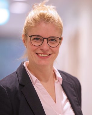 Eine Frau mit blonden Haaren, die zu einem Dutt gebunden sind, trägt eine Brille und einen dunklen Blazer über einem hellen Hemd. Sie lächelt freundlich in die Kamera. Der Hintergrund ist unscharf und hell.