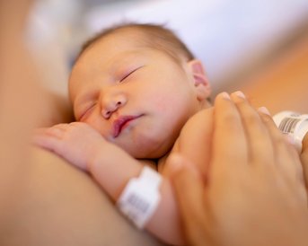 Ein neugeborenes Baby schläft friedlich auf der Brust einer Person, die eine Hand sanft auf dem Rücken des Babys hält. Das Baby trägt ein Identifikationsarmband.