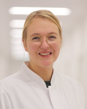 Eine lächelnde Frau trägt einen weißen Kittel und eine Brille. Sie steht in einem hellen, modernen Raum mit unscharfem Hintergrund. Ihr Gesicht zeigt Freundlichkeit und Professionalität.