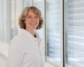 Eine lächelnde Frau in einem weißen Kittel steht neben großen Fenstern. Sie hat lockige Haare und blickt zur Kamera.