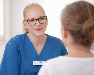 Eine medizinische Fachkraft in blauer Uniform spricht mit einer Patientin, die ihr den Rücken zuwendet.