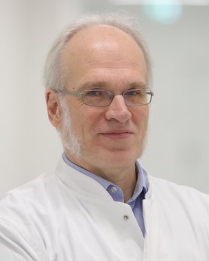 Ein Arzt mit Brille und grauen Haaren trägt einen weißen Kittel. Er steht mit verschränkten Armen in einem hellen, modernen Flur. Der Hintergrund ist unscharf und minimalistisch gestaltet.