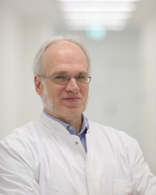 Ein Arzt mit Brille und grauen Haaren trägt einen weißen Kittel. Er steht mit verschränkten Armen in einem hellen, modernen Flur. Der Hintergrund ist unscharf und minimalistisch gestaltet.