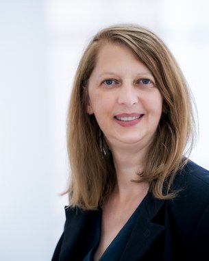 Eine Frau mit langen, glatten Haaren und einem schwarzen Blazer lächelt in die Kamera. Der Hintergrund ist hell und unscharf.