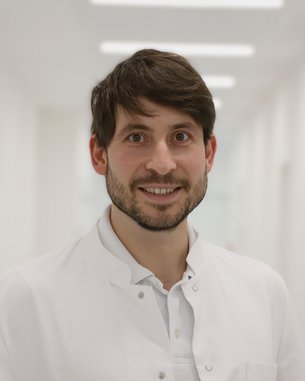 Ein junger Mann mit Bart trägt einen weißen Kittel und lächelt freundlich in einem hellen Flur.