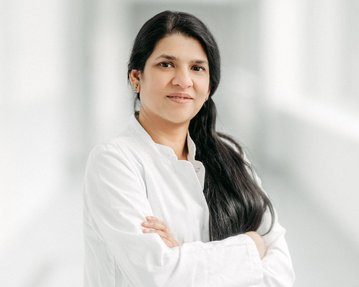 Eine Frau in einem weißen Kittel steht mit verschränkten Armen in einem hellen Flur. Sie hat lange, schwarze Haare und schaut direkt in die Kamera.