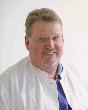 Ein Mann mit Brille trägt einen weißen Kittel und ein blaues Hemd. Er lächelt freundlich in die Kamera. Der Hintergrund ist hell und neutral gehalten.