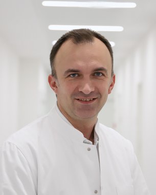 Ein Arzt in einem weißen Kittel lächelt freundlich in einem hellen Flur eines Krankenhauses.