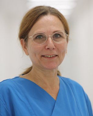 Eine Frau mit Brille trägt einen blauen medizinischen Kittel. Sie steht in einem hellen Flur und lächelt leicht. Ihr Haar ist zu einem Pferdeschwanz gebunden.