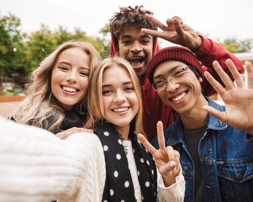 Vier junge Erwachsene posieren für ein Selfie, lächeln und zeigen Peace-Zeichen. Sie tragen lässige Kleidung und stehen in einer grünen Umgebung.