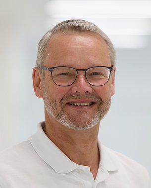 Ein Mann mit Brille und grauem Bart trägt ein weißes Polohemd und lächelt freundlich in einem hellen, modernen Raum.