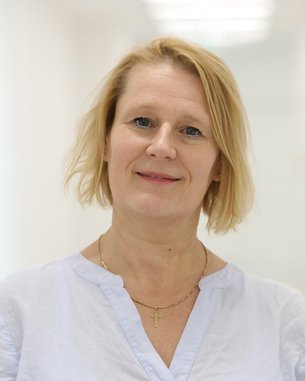 Eine Frau mit blonden, schulterlangen Haaren trägt ein hellblaues Oberteil und steht in einem hellen, modernen Raum.