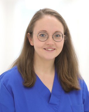 Eine Person mit langen, braunen Haaren trägt ein blaues medizinisches Outfit und runde Brille. Sie lächelt freundlich in die Kamera, vor einem hellen, neutralen Hintergrund.