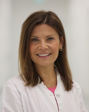 Chefärztin Emilia Wozniak Eine lächelnde Frau in einem weißen Kittel steht in einem hellen, modernen Flur. Sie hat lange, braune Haare und trägt goldene Ohrringe.