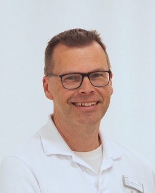 Ein Mann mit Brille und einem weißen Hemd lächelt freundlich in die Kamera. Der Hintergrund ist hell und neutral.