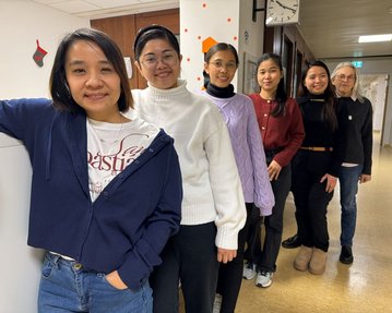 Sechs Personen stehen in einer Reihe in einem Flur. Sie tragen verschiedene Kleidungsstile und lächeln in die Kamera.