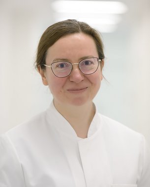 Dr. med Iea Grosul Eine Person mit Brille und einem weißen Kittel lächelt freundlich in einem hellen, neutralen Raum.