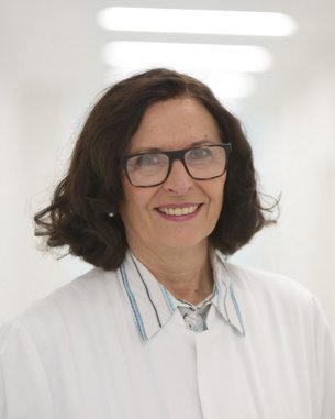 Eine Frau mit lockigem, braunem Haar trägt eine Brille und einen weißen Kittel. Sie lächelt freundlich und steht in einem hellen, modernen Raum. Der Fokus liegt auf ihrem Gesicht und ihrer professionellen Erscheinung.