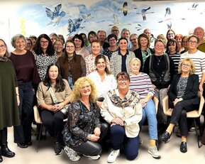 Eine Gruppe von etwa 50 Personen steht und sitzt in einem Raum, umgeben von einer Wand mit Vogelmotiven.