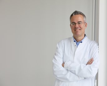 Ein Arzt in einem weißen Kittel steht mit verschränkten Armen an einer Wand und lächelt.