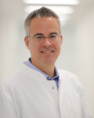 Ein Mann in einem weißen Kittel mit Brille lächelt freundlich in einem hellen, modernen Raum.