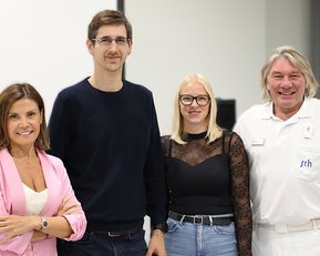 Vier Personen stehen zusammen in einem modernen Raum. Sie lächeln und tragen unterschiedliche Kleidung.