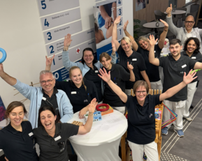 Eine Gruppe von Mitarbeitern steht in einem Empfangsbereich, lächelt und hebt die Hände. Sie tragen einheitliche schwarze Shirts und weiße Hosen.