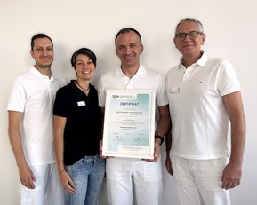 Vier Personen in weißen Outfits halten ein Zertifikat mit der Aufschrift "ZERTIFIKAT". Sie stehen vor einer neutralen Wand.