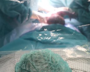 Eine Person in einem Operationssaal trägt eine grüne Haube und blickt auf einen chirurgischen Eingriff, der hinter einer transparenten Abdeckung stattfindet.