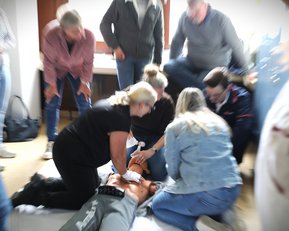 Mehrere Personen führen Erste-Hilfe-Maßnahmen an einer liegenden Person durch, während andere zuschauen und Anweisungen geben.