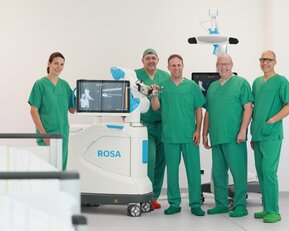 Fünf medizinische Fachkräfte in grüner Kleidung stehen um einen chirurgischen Roboter namens ROSA in einem modernen Operationssaal.
