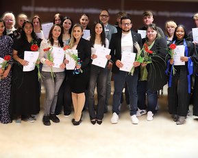 Gruppe von Absolventen hält Urkunden und Blumen, umgeben von Freunden und Familie, bei einer Feier zur Graduierung.