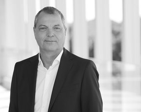 Mann in Anzug steht vor einer modernen Architektur mit Säulen. Er hat kurzes, graues Haar und lächelt leicht.