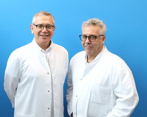 Zwei Männer in weißen Laborkitteln stehen vor einem blauen Hintergrund. Beide tragen Brillen und lächeln leicht.