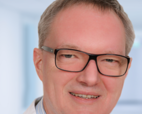 Ein Mann mit Brille trägt einen weißen Kittel und ein gestreiftes Hemd. Er lächelt freundlich in einem hellen, neutralen Hintergrund.