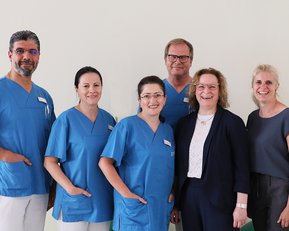 Sechs Personen in medizinischer Kleidung stehen zusammen, lächelnd. Drei tragen blaue Scrubs, während die anderen in formeller Kleidung gekleidet sind.