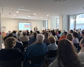 Ein Referent steht vor einem Publikum in einem modernen Raum, während er eine Präsentation auf einem Bildschirm zeigt.