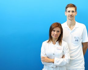 Zwei medizinische Fachkräfte stehen vor einem blauen Hintergrund. Die Frau trägt einen weißen Kittel, der Mann ein weißes Polohemd.