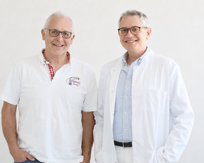 Zwei Männer stehen nebeneinander, einer trägt ein weißes Hemd und der andere einen weißen Laborkittel. Beide lächeln freundlich.