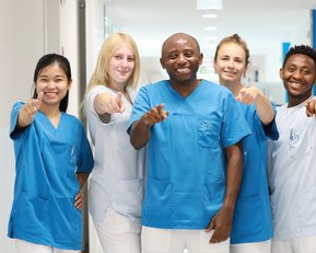 Sechs Personen in blauen Arbeitskleidung stehen in einem Flur und zeigen mit dem Finger auf die Kamera.