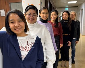 Sechs Personen stehen in einer Reihe in einem Flur, lächelnd und in unterschiedlichen Kleidungsstilen. Ein Wanduhren ist im Hintergrund sichtbar.