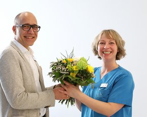 Ein Mann überreicht einer Frau mit blauer Uniform einen Blumenstrauß. Beide lächeln und stehen in einem hellen Raum.