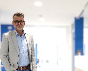 Ein Mann mit Brille und grauem Anzug steht in einem modernen Büro mit hellem Hintergrund.