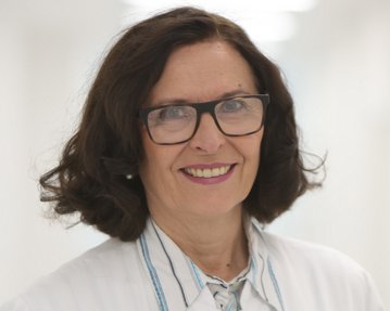 Eine Frau mit lockigem Haar und Brille trägt einen weißen Kittel. Sie lächelt freundlich in einem hellen, modernen Raum.